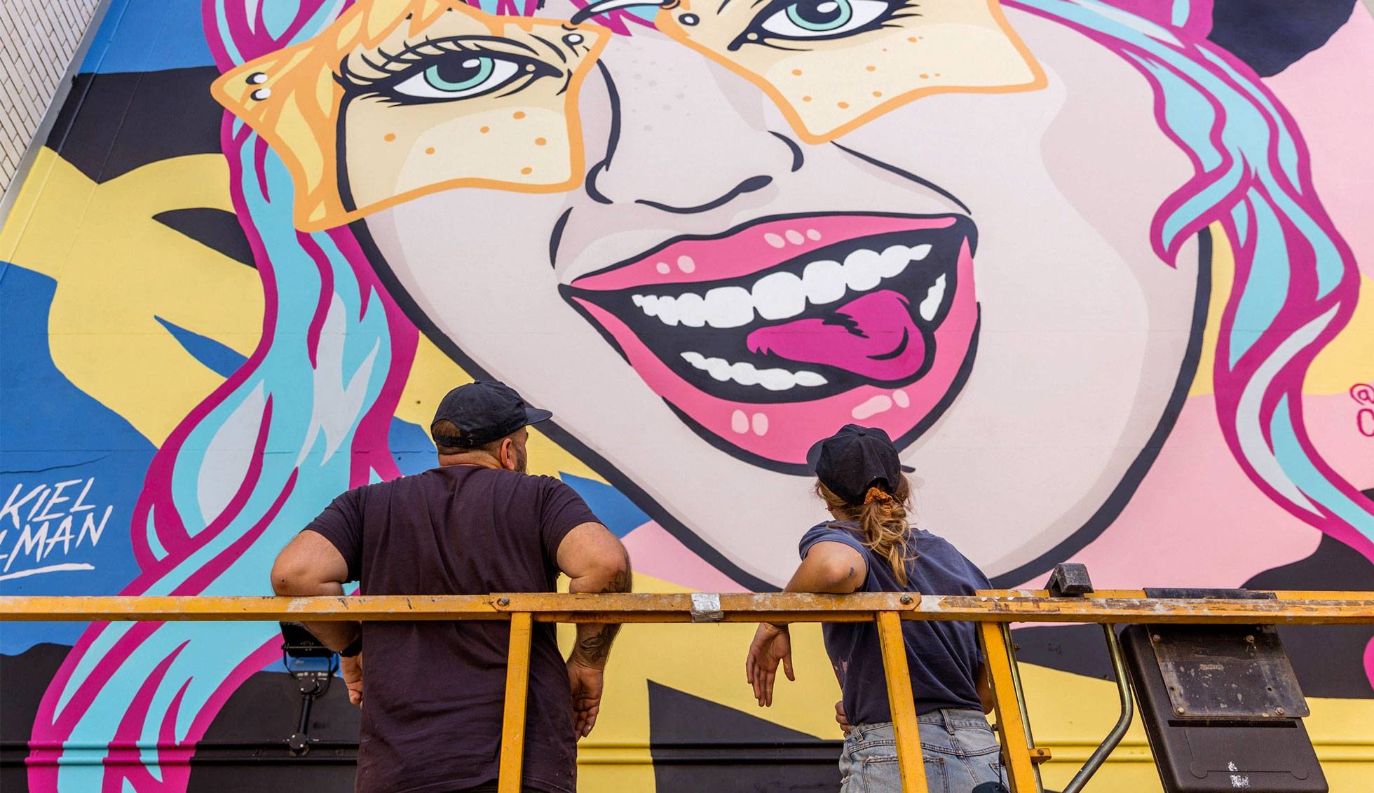 Two team members of Tillman Creative Co stand with their backs to the camera looking up at the large-scale mural they have just completed