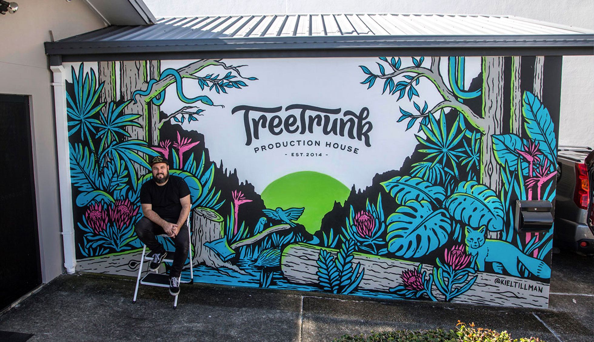Mural artist Kiel Tillman sits infront of his finished mural featuring the Treetrunk logo and a vibrant jungle scene
