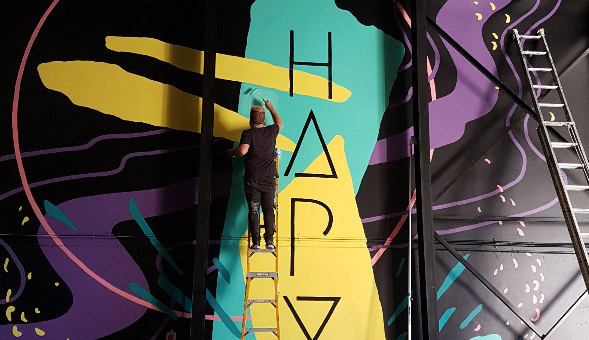 Gold Coast mural artist Kiel Tillman stands on a ladder painting a large-scale indoor mural