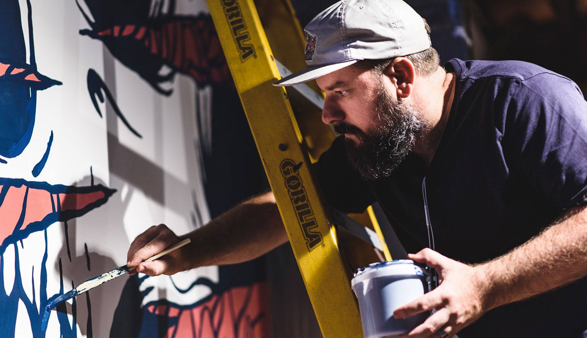 Kiel paints an indoor mural on a ladder, backlit by bright lights