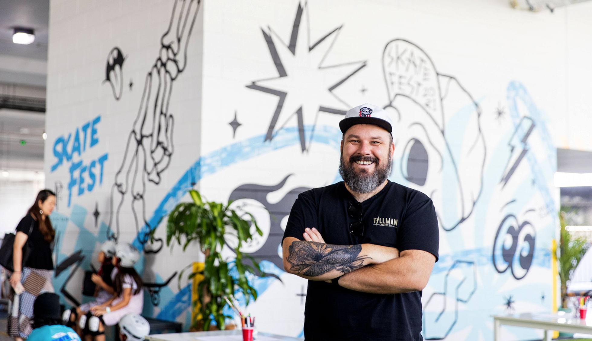 Mural artist Kiel Tillman stands smiling infront of his finished mural piece for Skatefest