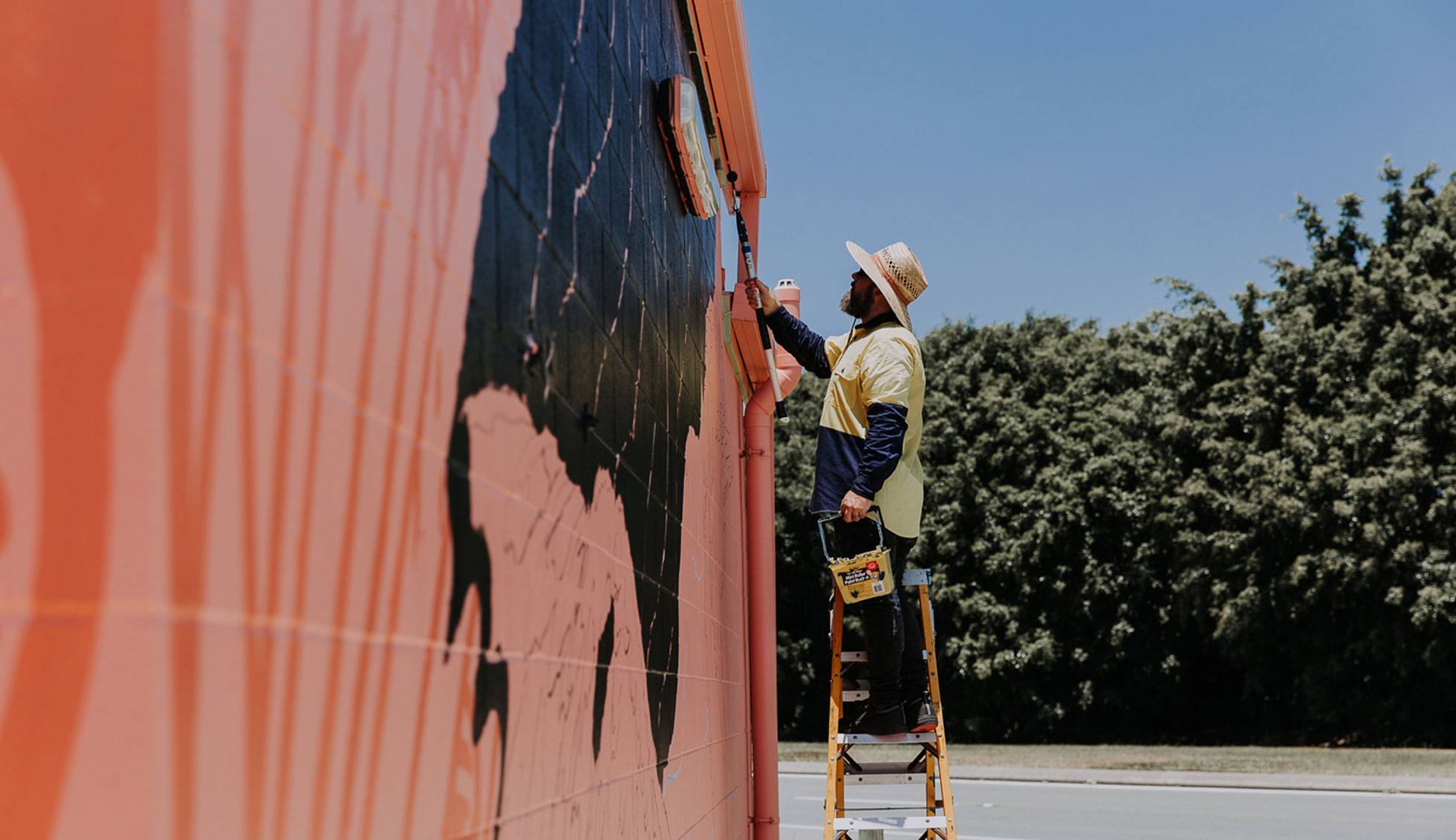 A mural artist paints an outdoor mural on a public wall. He paints a bird local to the area to make a meaningful art piece to residents.