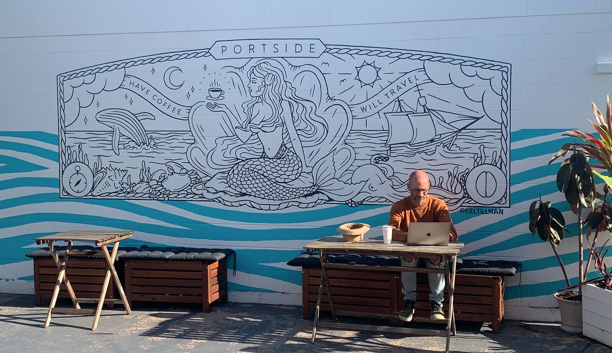 A man enjoys his coffee infront of Portside's outdoor mural, which features a mermaid, ships, ocean and the sun