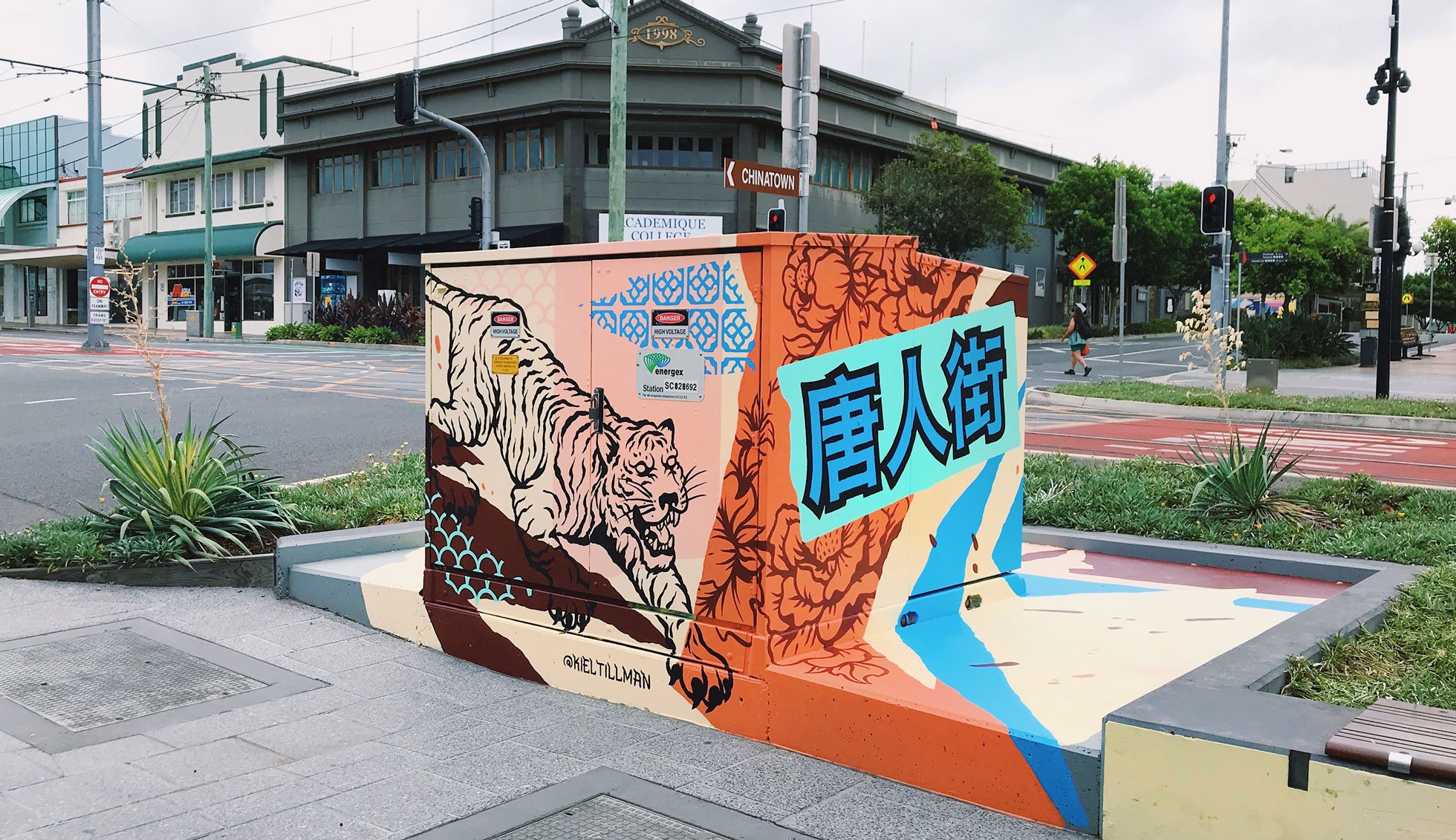 A public electrical box station covered in bright and colourful mural art relevant to the Chinatown era and culture. It features a tiger, Chinese glyphs and flower patterns. 